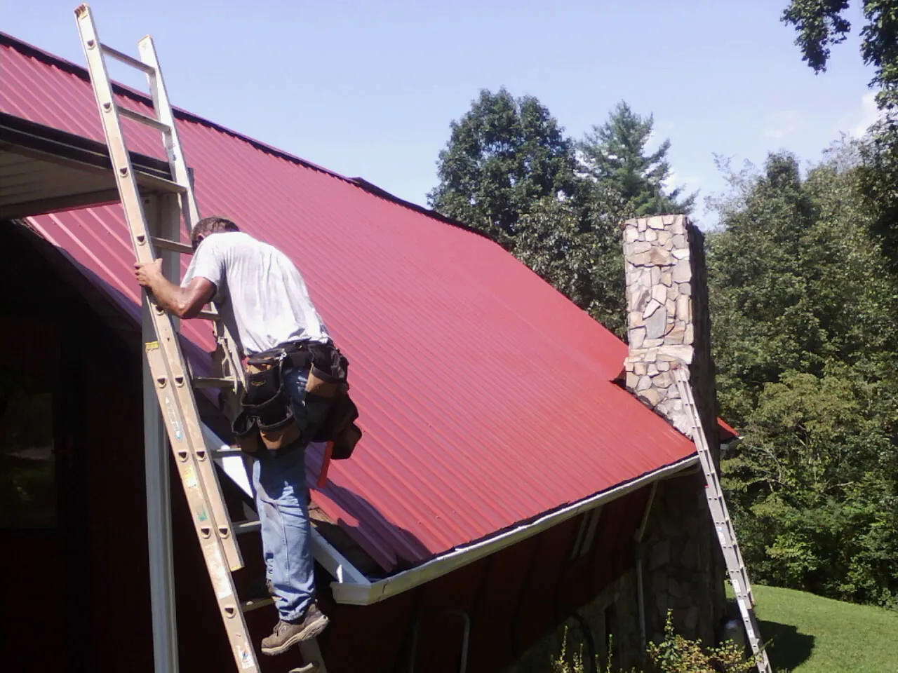 Roofer on a ladder beside a red metal roof and stone chimney — Soffit & Fascia Repair by Roofing Brothers in Marion